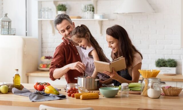 Happy parents helping daughter to cook. Mother with cookbook in hand, father with child on hand watching girl trying to catch something in pan. Family recreation and fun, loving relationships (Happy parents helping daughter to cook. Mother with cookbo